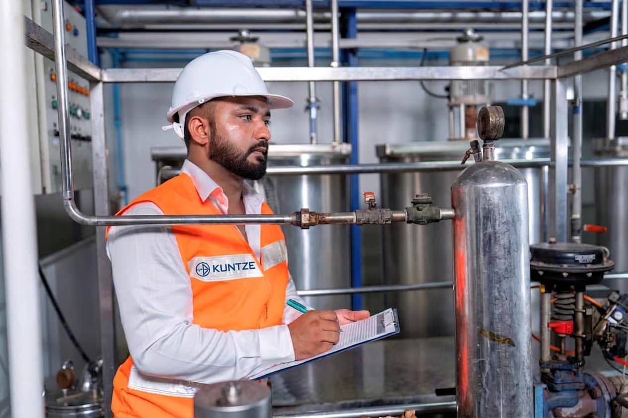 Engineer in a hard hat and high-visibility vest labeled “Kuntze” inspecting industrial piping and gauges while recording data on a clipboard in a water treatment or processing facility.